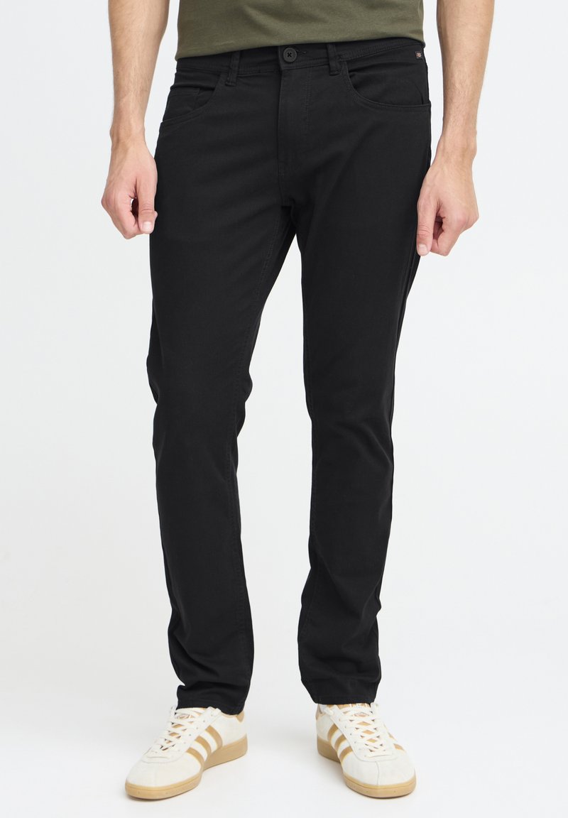 Pantalon en denim noir à coupe slim, avec cinq poches et une fermeture à boutons. Porté avec des baskets blanches à rayures beiges.