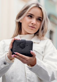 Černá kožená peněženka s vyvýšenými kruhovými vzory, uzavřená zaobleným patentem, držena v rukou na světlém pozadí.