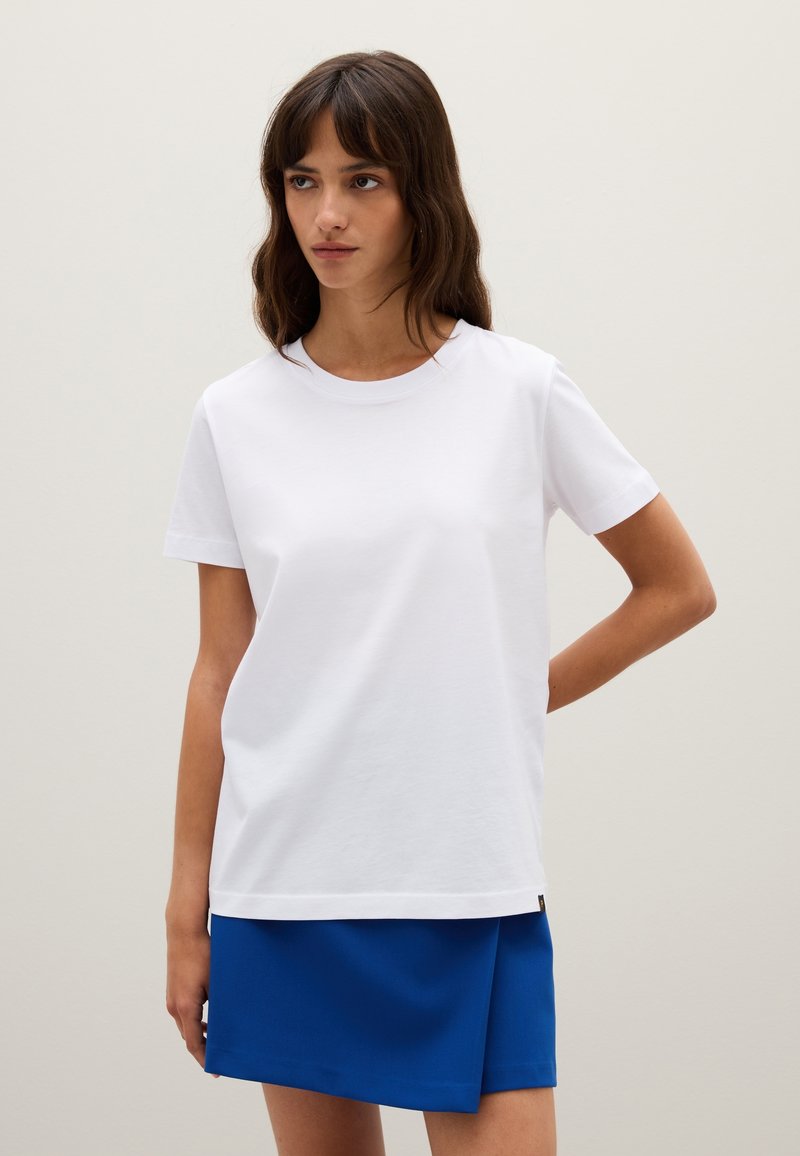 Frau mit braunen Haaren, die ein schlichtes weißes T-Shirt mit kurzen Ärmeln und einen blauen Rock trägt, vor einem beigen Hintergrund und nach links schaut.