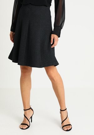 Vrouw draagt een zwarte, knielange, gestructureerde rok, een doorschijnende zwarte blouse met lange mouwen en zwarte hoge hakken met bandjes.