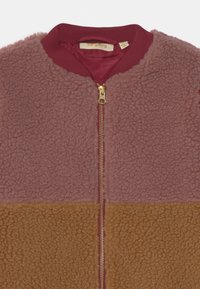 Giacca in pile con colletto bordeaux e cerniera dorata. Presenta una sezione superiore rosa e una sezione inferiore marrone texturizzata. Tessuto morbido.