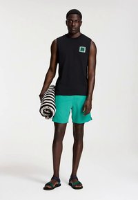 Black sleeveless shirt, green swim shorts, black sandals; holding a striped towel. Simple design, contrasting colors, and casual fit.