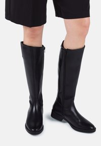 Black knee-high leather boots with side zippers worn with black shorts on visible legs against white background.