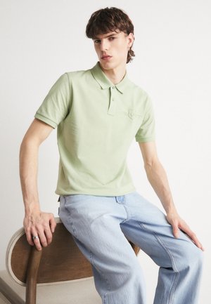 Camisa polo verde claro con cuello y mangas cortas, con un logo en relieve. Combinada con jeans azul claro, sentado en una silla de madera.