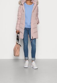Gepolsterter rosa Mantel mit einer fellgefütterten Kapuze, hellblauer Pullover, Jeans und weiße Turnschuhe. Trägt eine weiche, beigefarbene Handtasche.