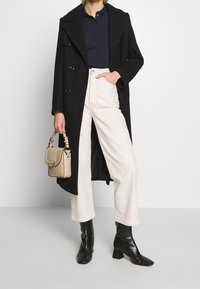 Modell i en svart kappa och marinblå polo håller en beige handväska, iförd vita vida byxor och svarta ankelskor, mot en neutral bakgrund.