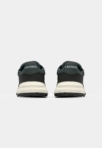 Black athletic shoes with a textured upper, green accents, and white midsole. Features "LACOSTE" branding on the heel and a durable rubber outsole.