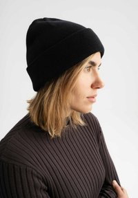 Zwarte geribbelde brei beanie met een opgerolde rand, gedragen door een persoon met gelaagd haar, gecombineerd met een donkere geribbelde trui. Zijkant profielweergave.