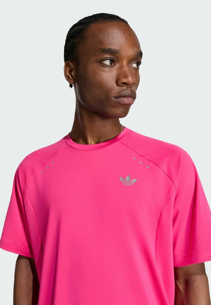 Hombre con cabello trenzado que lleva una camiseta rosa brillante de Adidas mirando hacia un lado sobre un fondo claro y liso.