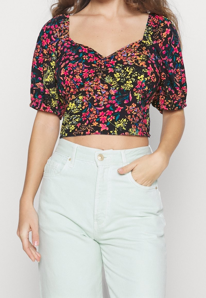 Květinový crop top v černé barvě s živými vzory v růžové, žluté a zelené; krátké nafouknuté rukávy a výstřih do V. Vzhledem s lehce mátovými džíny.