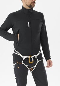 Homme portant une veste noire zippée et un harnais d'escalade avec un mousqueton sur fond blanc, la main dans la poche.