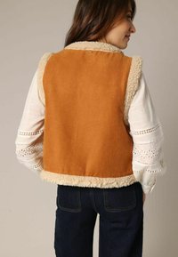 Gilet en faux suède brun avec une bordure en fausse fourrure crème, design sans manches et texture lisse, porté sur une chemise blanche à manches longues brodée.