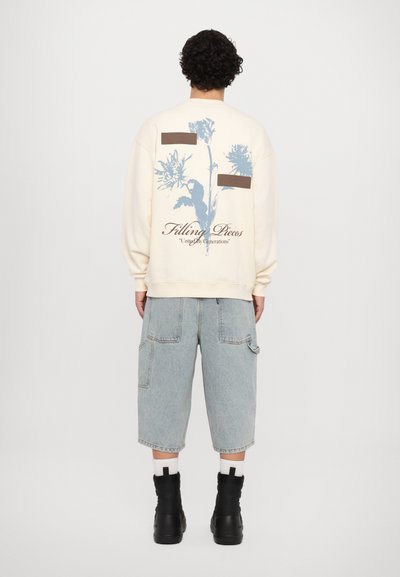 Cremefarbenes Sweatshirt mit blauem Blumenmuster und braunen Patches auf der Rückseite, kombiniert mit hellblauen Cargo-Shorts und schwarzen Knöchelboots.