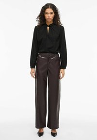 Svart teksturert bluse med åpning i nøkkelhullstil, kombinert med høytlivede, vide burgunderfargede bukser i kunstlær og spisse svarte hæler.