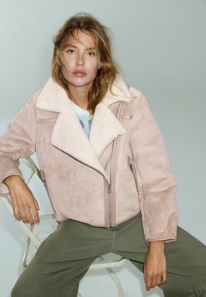 Femme aux cheveux ébouriffés portant une veste en peau lainée rose clair et un pantalon vert, assise sur une chaise blanche devant un fond uni.