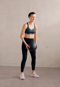 Sportowy biustonosz i legginsy Nike w ciemnych odcieniach. Osoba trzyma czarną piłkę lekarską. Jasne buty z białymi akcentami. Minimalistyczne tło.