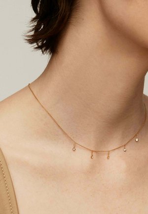Collar de oro con una delicada cadena y cinco pequeños charms colgantes. Presenta finos eslabones y una textura suave, descansando sobre un cuello de tono de piel.