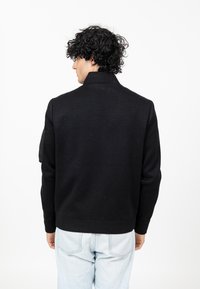 Zwarte gebreide jas met een hoge kraag, geribbelde manchetten en zoom. Kenmerkt zich door een patch op de linker mouw. Draagt bij lichtblauwe jeans.