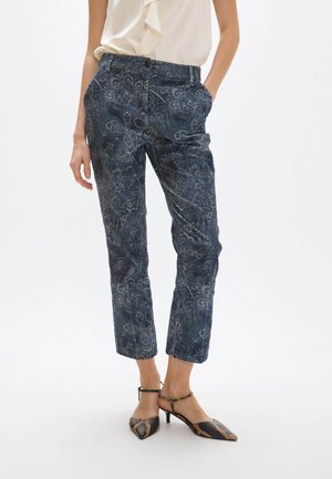 Mujer con pantalones cortos azul oscuro con estampado de paisley y tacones slingback con punta en estampado de animal, de pie sobre fondo blanco.