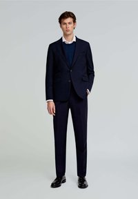 Blazer marino con dos botones y bolsillos, superpuesto a un suéter oscuro. Pantalones ajustados en marina a juego. Zapatos negros.