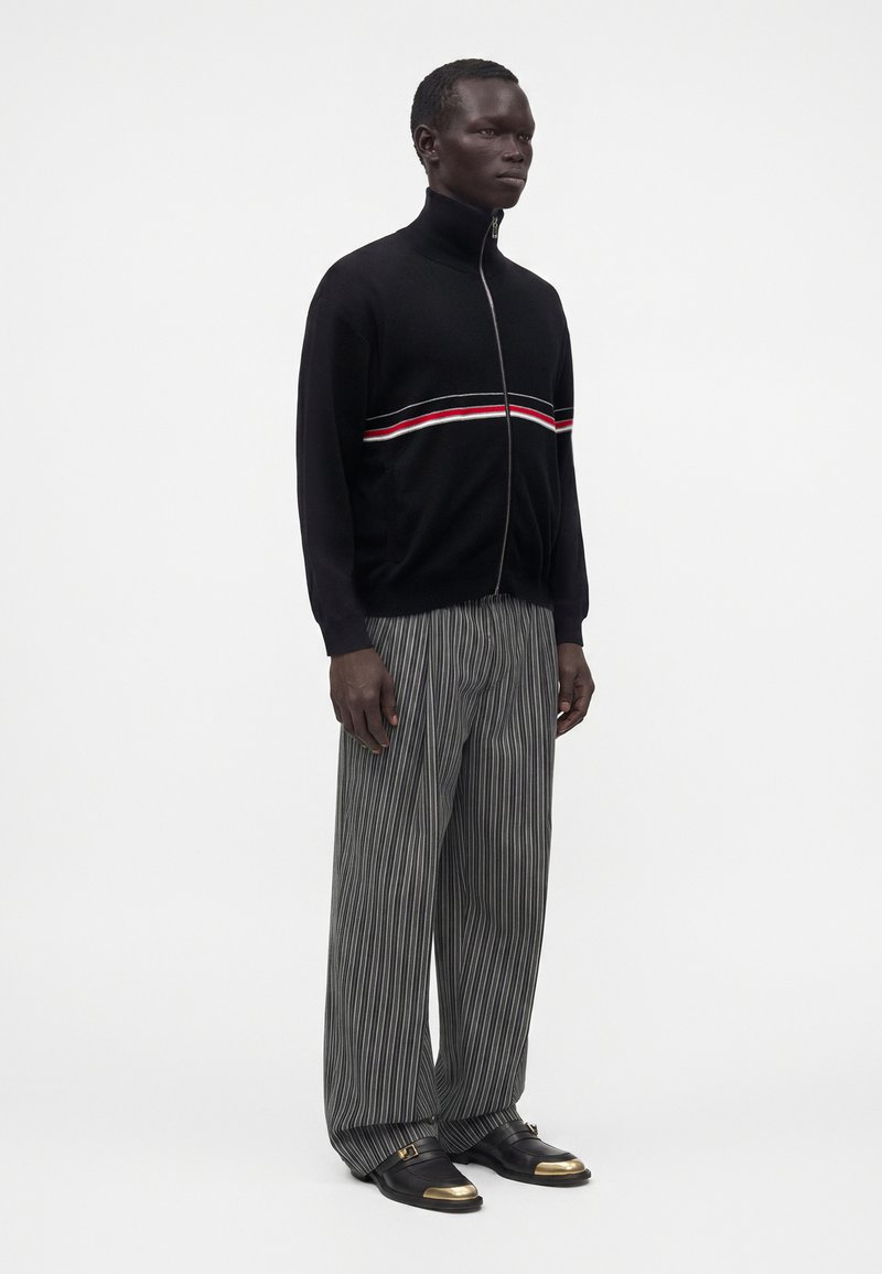 Homme portant un pull noir zippé avec une bande rouge et blanche, un pantalon large gris à fines rayures, et des chaussures noires avec des embouts dorés.