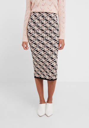 Vrouw draagt een potloodrok met zwart, roze en wit geometrisch patroon, lichtroze gebreide trui en witte puntige schoenen.