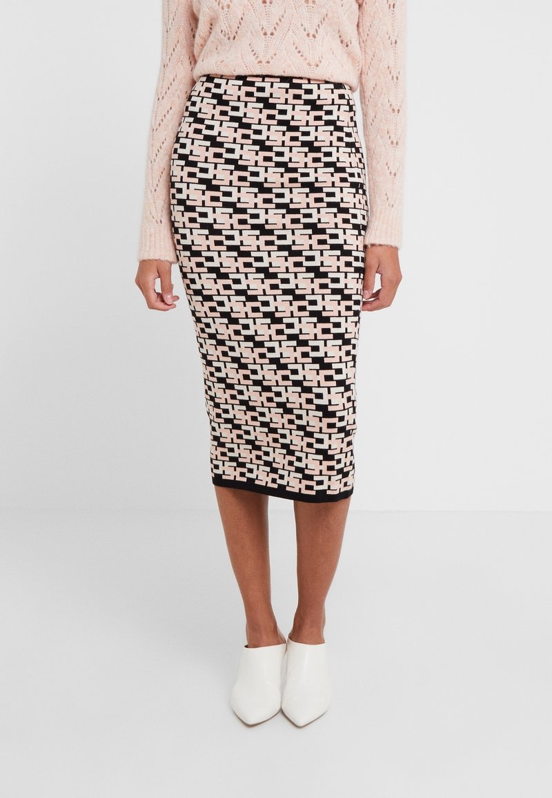 Femme portant une jupe crayon à motifs géométriques noirs, roses et blancs, un pull en maille rose clair et des chaussures blanches à bouts pointus.