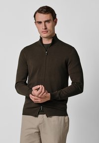 Brązowy sweter zapinany na zamek wykonany z gładkiego dzianinowego materiału. Posiada wysoki kołnierz i dopasowane rękawy, noszony na ciemnej koszuli.