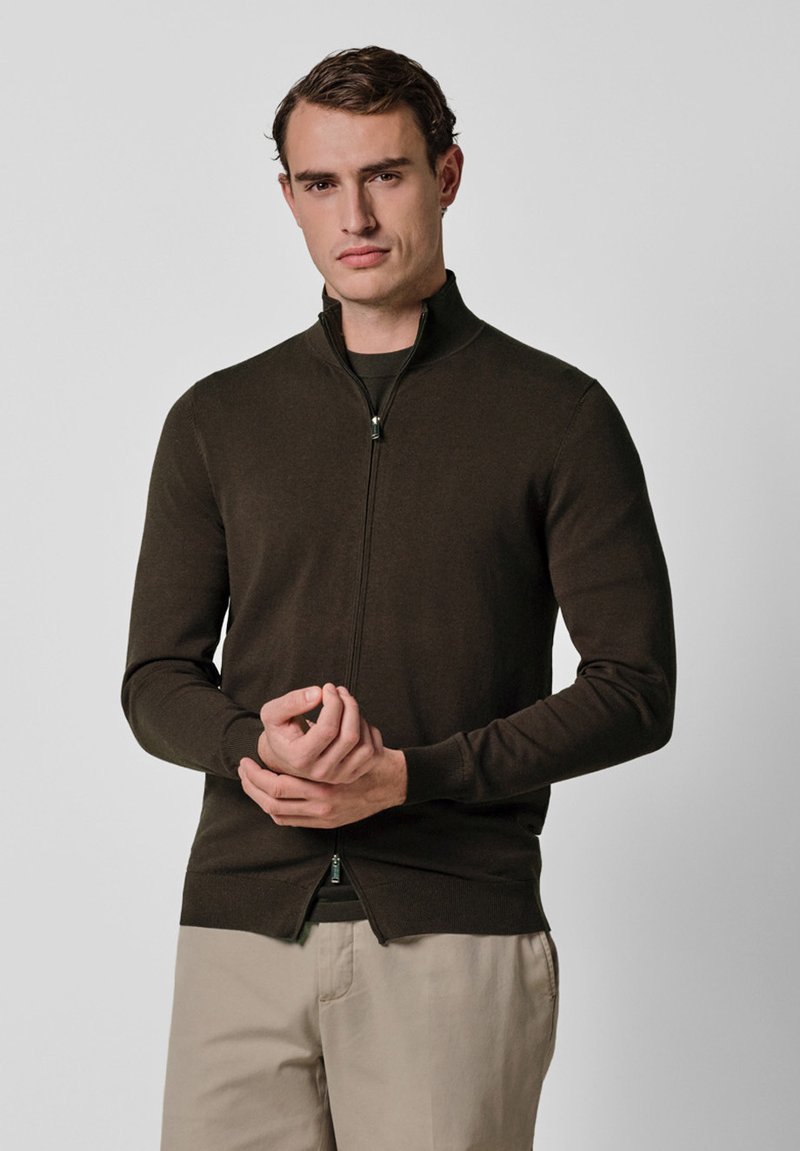 Brązowy sweter zapinany na zamek wykonany z gładkiego dzianinowego materiału. Posiada wysoki kołnierz i dopasowane rękawy, noszony na ciemnej koszuli.