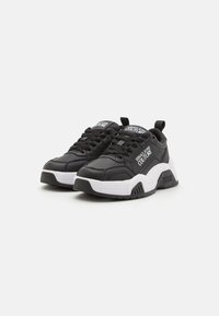 Zapatillas negras con una suela blanca gruesa. Fabricadas en material sintético suave, con diseño de cordones y acentos de logo en la lengüeta y el talón.