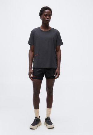 Fiatal személy rövid, göndör hajjal, fekete sportos pólót és rövidnadrágot visel, bézs színű zokniban és fekete sportcipőben áll egy egyszínű háttér előtt.