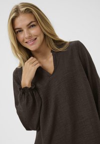 Dunkelbraune Bluse mit V-Ausschnitt und weichem, strukturiertem Stoff. Verfügt über lockere, geraffte Ärmel für eine entspannte Passform. Einfaches und elegantes Design.