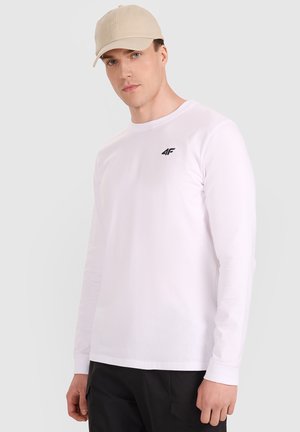 Man wearing beige cap, white long-sleeve shirt with black logo on chest, and black pants, standing against a light gray background.