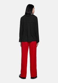 Schwarzes Langarmshirt mit einer Rückenfalte, kombiniert mit weiten roten Hosen mit seitlichen Schlitzen und schwarzen Schuhen mit klobigen Sohlen.