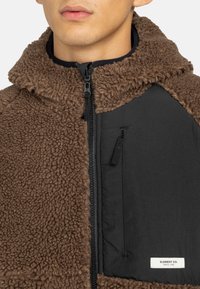 Muže s hnědou fleece bundou sherpa, která má černou zipovou kapsu na hrudi a štítek Element Co. na kapse.