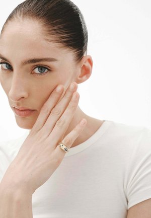 Anillo de oro con piedras azules y transparentes, con un diseño suave y redondeado. La mano del modelo reposa sobre su rostro, mostrando el accesorio.