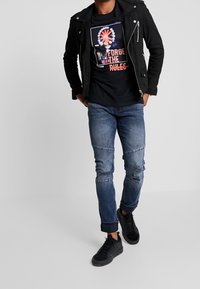 Man som bär en svart jacka, svart grafisk T-shirt med texten "Forge the Rules", slitna blå jeans och svarta sneakers, står mot en grå bakgrund.