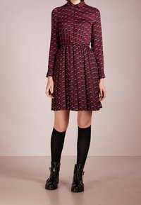 Abito lungo bordeaux a maniche lunghe con vita aderente, caratterizzato da un motivo a luna crescente ripetuto e gonna plissettata, abbinato a calze nere fino al ginocchio.