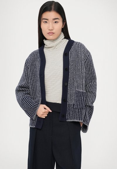 Pletený kardigán v tmavomodrej farbe s ľahkými pruhmi, s rebrovanou štruktúrou, zapínaním na gombíky a predným vreckom. Pod ním rolák.