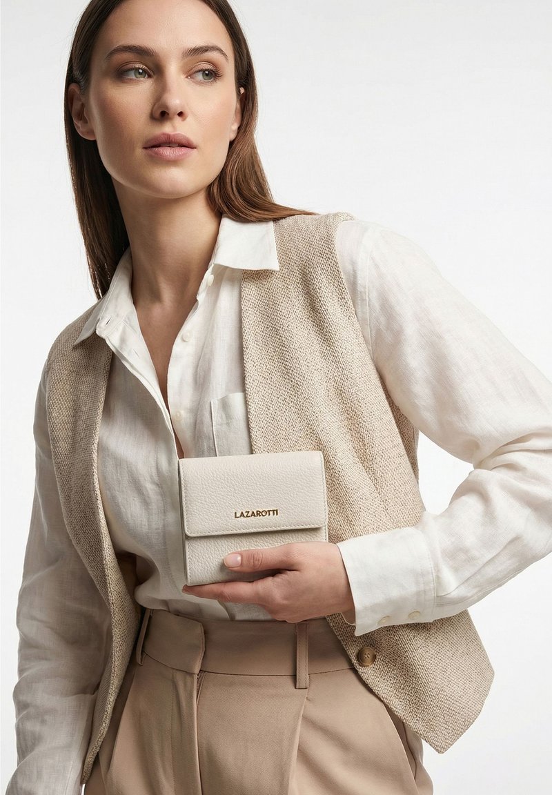 Frau trägt eine beige Weste über einem weißen Hemd und einer Hose, hält eine kleine weiße Geldbörse mit in Gold geprägtem "LAZAROTTI".