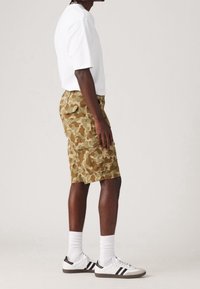 Pantalones cortos de camuflaje en tonos marrón y beige, hechos de un tejido ligero, con múltiples bolsillos, combinados con una camiseta blanca y zapatillas.