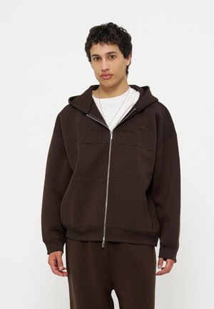 Młody mężczyzna ubrany w ciemnobrązową bluzę z zamkiem na białą koszulę, stojący na jasnym, jednolitym tle, zwrócony twarzą do przodu z neutralnym wyrazem.