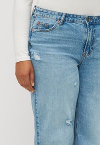 Light blue denim jeans with distressed detailing, visible stitching, a high waist, and copper button accents. Worn with a white long-sleeve shirt.