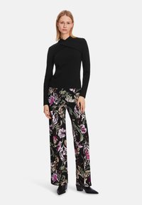 Schwarzes Langarmoberteil mit gedrehter Frontgestaltung, kombiniert mit weiten schwarzen Hosen mit bunten Blumenmustern. Schwarze Absatzstiefeletten.