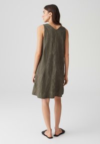 Sleeveless olive green dress with a textured leaf pattern, a V-shaped back, and an A-line silhouette. Worn with black slide sandals.