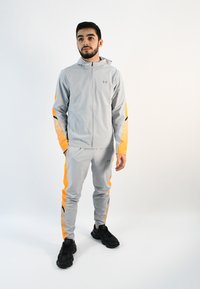 Chándal atlético gris claro con acentos naranjas, que incluye una chaqueta con cremallera y pantalones ajustados con detalles en negro y una textura suave.