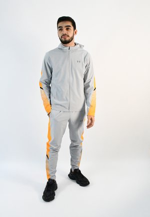 Chándal atlético gris claro con acentos naranjas, que incluye una chaqueta con cremallera y pantalones ajustados con detalles en negro y una textura suave.