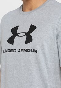 Grå bomullströja med en stor svart Under Armour-logga och text under i fet, kontrasterande svart font. Kort ärm.
