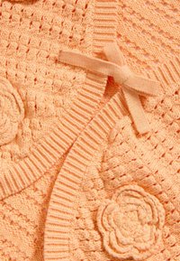 Gilet en tricot pêche avec un motif texturé, des bords côtelés et des appliqués floraux. Inclut un accent en nœud. Matière douce et légère.