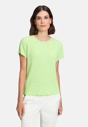 Haut court à manches courtes texturé vert clair avec un col rond et une coupe ample. Tissu à motif gaufré subtil, associé à un pantalon blanc.